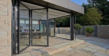 Glasfalttüren öffnen sich in einem modernen Raum mit Steinmauern im Hintergrund, Terrassenplatten auf dem Boden und einem Tisch samt Stühlen im Inneren des Gebäudes.