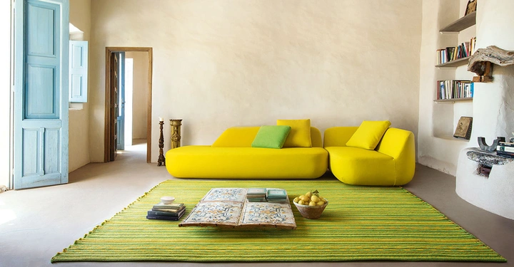 Gelbes Sofa mit Kissen steht in einem hellen Raum mit cremefarbenen Wänden liegendem gestreiften Teppich einem Stapel Bücher und einer Obstschale. Regal mit Büchern rechts blaues Fenster und Tür links.