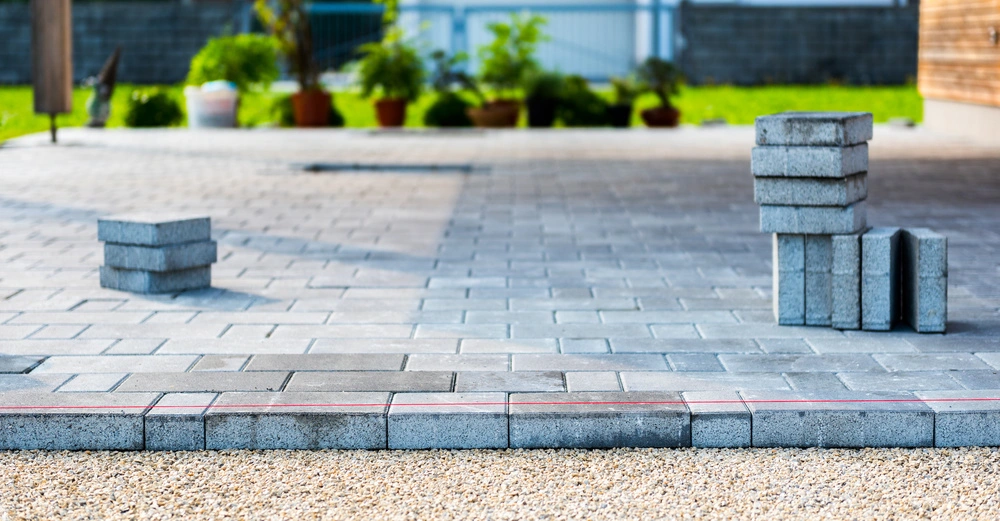 Stapel von Pflastersteinen auf einer teilweise fertiggestellten Palette vor einem Garten mit einem Haus im Hintergrund.
