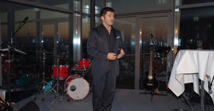 Galerie-Bild 64: Ein Mann steht auf einer Bühne und spricht in ein Mikrofon. Im Hintergrund befinden sich ein Schlagzeug und Gitarren. Die Bühne steht in einem modernen Raum mit Glaswänden.