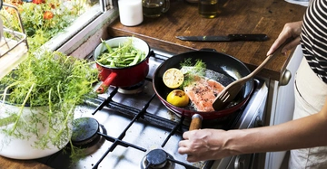 Person brät Lachs mit Zitrone in einer Pfanne auf einem Gasherd neben grünem Gemüse in einem Topf in einer Küche mit Pflanzen am Fenster.