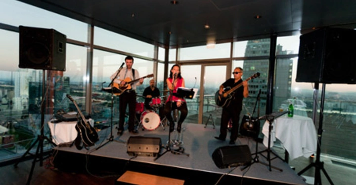 Galerie-Bild 47: Eine Band spielt auf einer kleinen Bühne in einem modernen Raum mit großen Fenstern. Im Hintergrund sind Hochhäuser sichtbar. Es gibt zwei Gitarristen, einen Schlagzeuger und eine Sängerin.