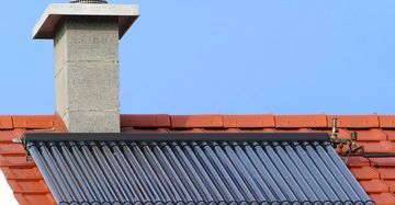 Solarkollektor ist auf einem roten Ziegeldach installiert mit einem grauen Schornstein im Hintergrund unter klarem blauem Himmel.