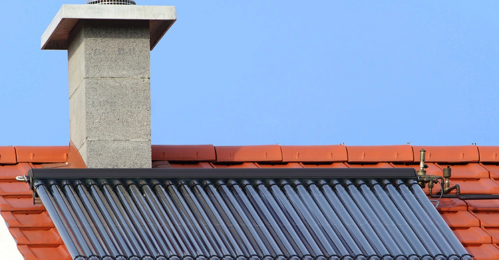 Solarkollektor ist auf einem roten Ziegeldach installiert mit einem grauen Schornstein im Hintergrund unter klarem blauem Himmel.