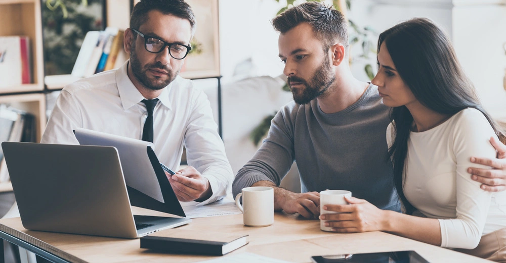 Mann mit Brille erklärt einem Paar Dokumente im Büro. Der Mann zeigt auf das Dokument, während das Paar aufmerksam zuhört. Beide Paare halten Tassen. Laptop und Tablet auf dem Tisch.