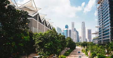 Mehrspurige Straße mit fahrenden Autos umgeben von modernen Hochhäusern und dichtem Laubwerk klare Himmel und vereinzelte weiße Wolken im Hintergrund.