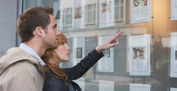 Ein Mann und eine Frau betrachten Immobilienanzeigen in einem Schaufenster in einer städtischen Umgebung. Die Frau zeigt mit dem Finger auf eine bestimmte Anzeige.