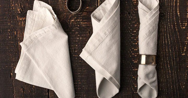 Drei cremefarbene, strukturierte Leinen-Servietten, in verschiedenen Stilen auf einem dunklen Holztisch gefaltet. Eine davon wird von einem silbernen Serviettenring gehalten.