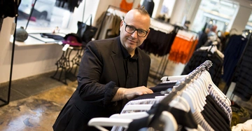 Ein Mann in schwarzer Kleidung durchsucht Kleidungsstücke an einem Kleiderständer in einem modernen Bekleidungsgeschäft mit großen Fenstern und unterschiedlichen Kleidungsstücken um ihn herum.