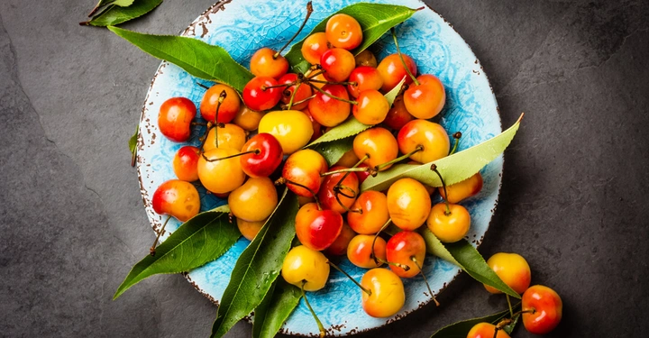 Mehrfarbige Kirschen und grüne Blätter liegend auf einem blau-weißen Teller, der auf einer dunklen Oberfläche platziert ist.