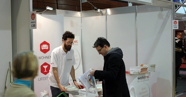 Galerie-Bild 23: Mann hinter einem Tisch in einem Messe-Stand gibt Informationsmaterial an einen anderen Mann. Im Hintergrund sind Werbeschilder von wohnnet. Text: wohnnet WO UND WIE WOHNEN Halle 6 130.