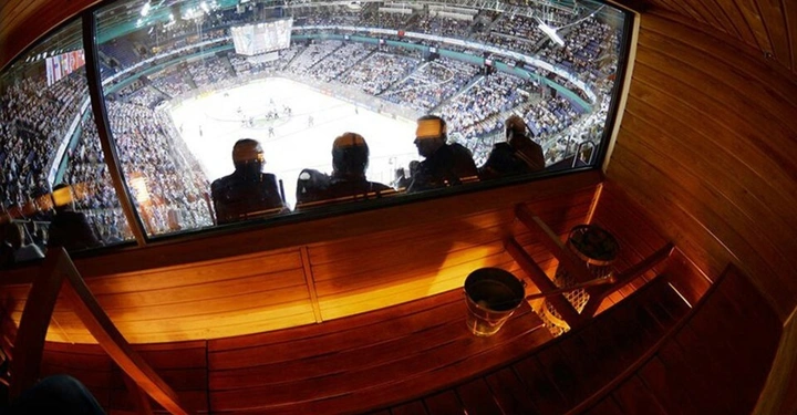 Saunagäste beobachten ein Eishockeyspiel in einem Stadion durch ein großes Fenster aus einer hölzernen Sauna heraus. Die Tribünen sind voll mit Zuschauern.