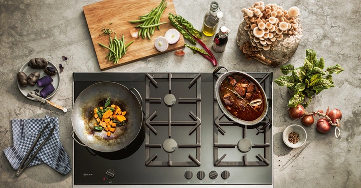 Pfanne mit Gemüse brät auf einem Herd vor bunten Zutaten auf einem Tisch wie Pilzen Bohnen Zwiebeln Kartoffeln Olivenöl und Gewürzen in einem organisierten Kochumfeld.