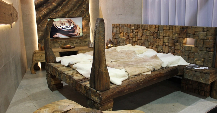 Galerie-Bild 3: Ein rustikales Holzbett mit Decken und Kissen steht in einem Schlafzimmer aus natürlichen Materialien. Im Hintergrund ist ein eingeschalteter Fernseher über einem kleinen Tisch zu sehen.