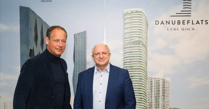 Zwei Männer stehen nebeneinander und lächeln in die Kamera im Hintergrund befinden sich hohe, moderne Gebäude. Text: EIN PROJEKT VON S+B GRUPPE SORAVIA DANUBEFLATS LEBE HOCH.