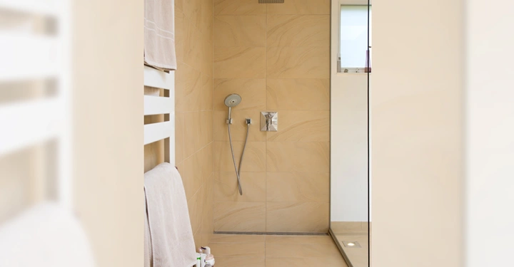 Galerie-Bild 2: Dusche an Wand montiert in einer geräumigen Duschkabine mit beigen Fliesen und einem Glasabtrennung Fenster rechts Handtücher und Duschprodukte auf einem weißen Handtuchhalter links