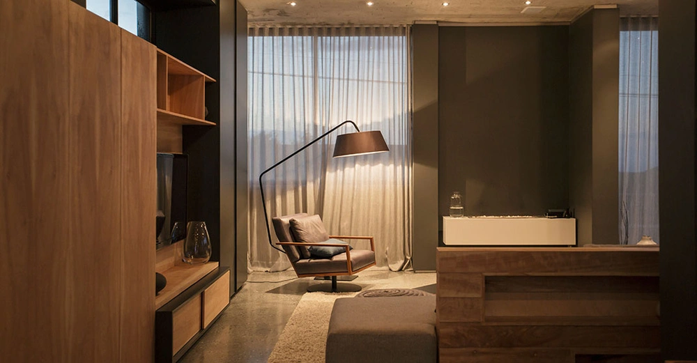 Modernes Wohnzimmer mit einer Wohnwand aus Holz und einem grauen Lesesessel, der von einer großen Stehlampe beleuchtet wird. Eingelassen Spots in der Sichtbetondecke. 