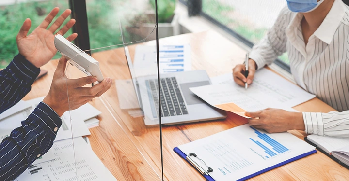 Eine Person zeigt ein Dokumentiersystem während einer Besprechung durch eine Plexiglasscheibe einem anderen, der Notizen auf Papier neben einem Laptop in einem Büro macht.