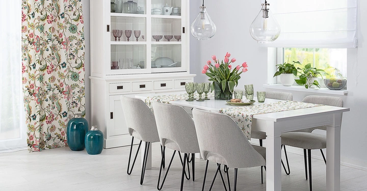 Weißer Esstisch mit sechs Stühlen dekoriert mit grünem Geschirr und rosa Tulpen in einem hellen Raum mit großem Fenster eine Anrichte und blumigen Vorhängen in lebendigen Farben.