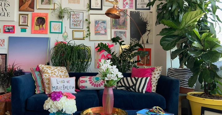 Blauer Sofa steht inmitten von Pflanzen und Kissen in einem bunt dekorierten Raum. Der Hintergrund zeigt eine Wand voller Kunstwerke und Rahmen, weiße und gelbe Blumen befinden sich auf einem rosafarbenen Tisch. Texte auf Bildern: Land of MAKEBELIEVE HERE’S TO THE CRAZY ONES