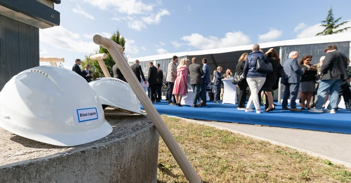 Helme liegen auf einem Betonpfosten mit Namensschild Blaue Lagune. Im Hintergrund stehen Menschen in formeller Kleidung auf blauem Teppich vor einem modernen Gebäude und unterhalten sich.