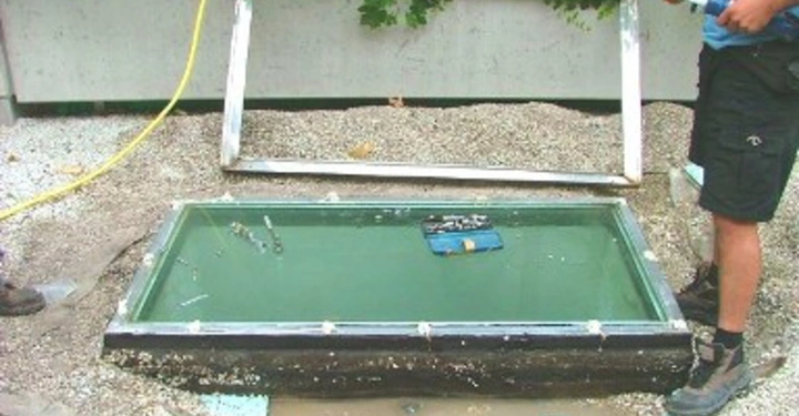 Galerie-Bild 31: Ein Rechteckfenster liegt auf dem Boden in einem kleinen Wasserbecken während zwei Personen werkzeugartige Gegenstände in der Nähe halten Es gibt Kieselsteine und Pflanzen im Hintergrund