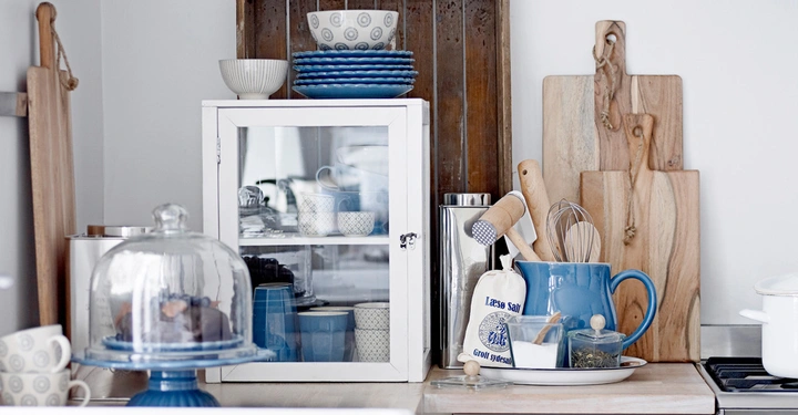 Galerie-Bild 6: Kuchen auf einem blau-weißen Teller unter einer Glasglocke in einer Küche mit einem weißen Schrank voller Geschirr Holzschneidebretter und blauen Utensilien auf der Arbeitsfläche Holzbrett im Hintergrund