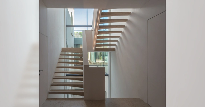 Freischwebende Treppe verbindet mehrere Stockwerke in einem modernen hellen Gebäude mit großen Fenstern durch die Tageslicht einfällt und einen Blick auf eine Grünfläche draußen bietet