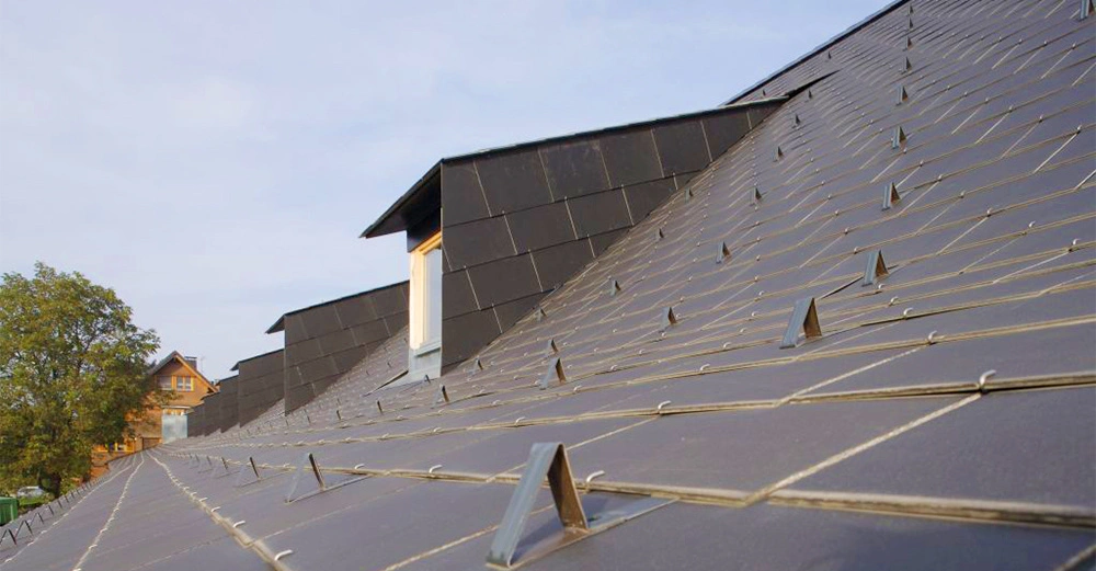 Dachfenster ragen aus einer großen geneigten Dachfläche heraus, die mit Solarpaneelen bedeckt ist. Im Hintergrund sind Bäume und ein weiteres Haus sichtbar.