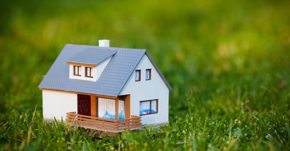 Ein weißes Modellhaus mit grauem Dach steht auf grünem Gras in einem Außenbereich