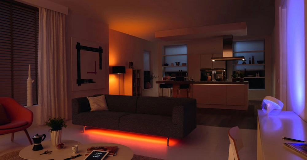 Graues Sofa leuchtet am Boden orange steht in einem modernen Wohnzimmer und blickt auf eine offene Küche mit eingebauter Beleuchtung. Tablets und Tassen liegen auf Tisch.