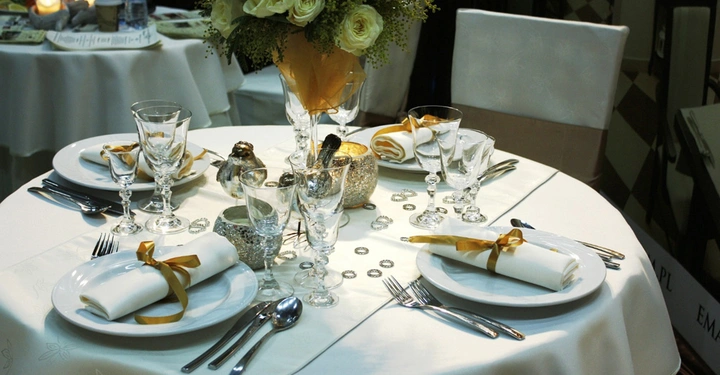 Rundtisch ist festlich gedeckt mit weißen Tellern Gläsern und goldenen Serviettenröllchen Im Zentrum steht ein Blumenstrauß mit weißen Blüten Im Hintergrund weitere gedeckte Tische