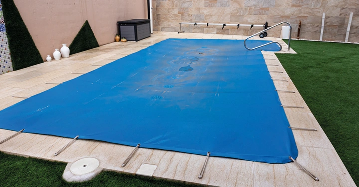 mit dunkelblauer Plane vollkommen abgedeckter Pool, Plane an Riemen befestigt. Poolumrandung mit hellbrauenen Pflastersteinen