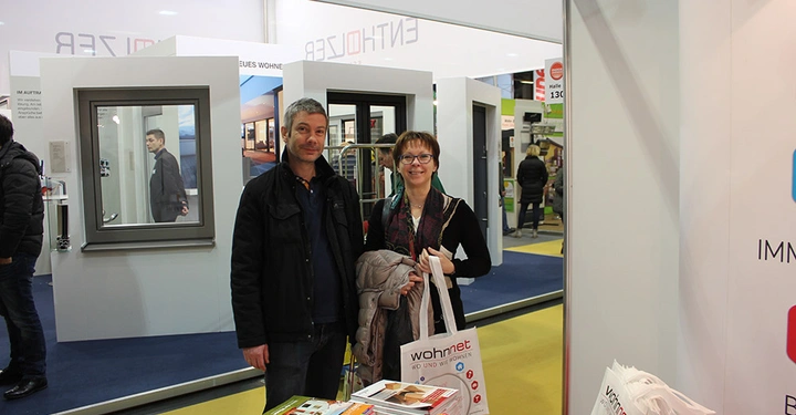 Galerie-Bild 14: Zwei Personen lächelnd stehend auf einer Messeumgebung haltend Taschen Beschriftungen darauf wohnnet red und fenster tür und Immobilien im Hintergrund Fenster und Türen Displays und anderer Messebesucher.