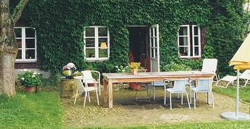Holztisch steht mit mehreren Stühlen auf einer Terrasse vor einem mit Efeu bewachsenen Haus umgeben von Blumen und Gartenmöbeln. Eine Tür zur Terrasse ist offen.