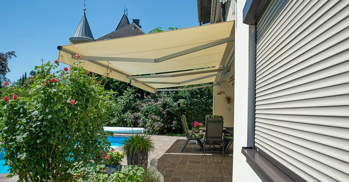Markise spendet Schatten über einer Terrasse neben einem Pool. Umliegende Rosensträucher und ein neugotisches Gebäude im Hintergrund vervollständigen die Szenerie an einem sonnigen Tag.