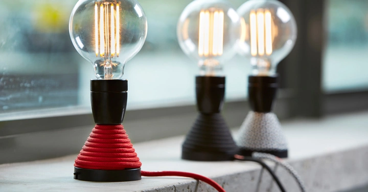 Galerie-Bild 5: Drei Glühbirnen leuchten still auf schwarzen Sockeln mit bunten Textilkabeln in Rot Schwarz und Weiß auf einer Fensterbank in einem modernen Raum.