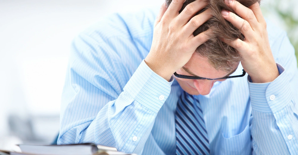 Mann hält Kopf in den Händen sitzt an einem Schreibtisch im Büro vor einem Stapel von Akten oder Papieren
