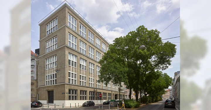Galerie-Bild 16: Ein mehrstöckiges Gebäude mit vielen Fenstern steht an einer Straßenecke große Bäume wachsen daneben geparkte und fahrende Autos befinden sich auf der Straße eine Person überquert die Straße.