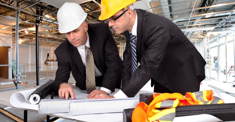 zwei Bauführer im Anzug mit Baustellenhelm begutachten Pläne in einer großen Halle, in der gebaut wird