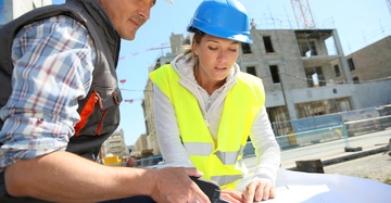 Mann und Frau mit Helm und Warnweste besprechen auf großer Baustelle den Bauplan, der ein Teil des Bauaktes ist. Im Hintergrund Rohbau eines mehrstöckigen Gebäudes