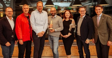 Sieben Personen stehend lächelnd innen in einem Raum mit Hängelampen eine Person hält eine Trophäe kontext ist moderne Büroeinrichtung im Hintergrund ist ein Besprechungsbereich erkennbar.