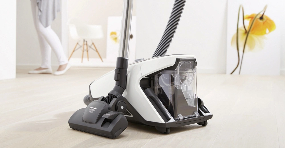 Staubsauger saugt auf hellem Holzboden; im Hintergrund weiße Wände mit gelbem Blumenbild und eine Person geht barfuß in weißen Leggings vorbei. Text Miele Blizzard CX1 PLUS.