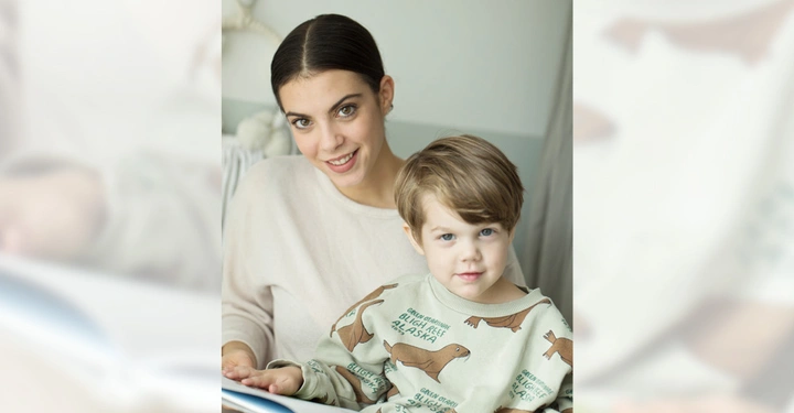 Galerie-Bild 4: Frau lächelt und hält Buch während sie mit einem Kleinkind in einem hellen Zimmer auf einem Stuhl sitzt beide schauen in die Kamera das Kleinkind trägt einen Pullover mit Tiermotiven