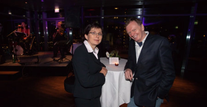 Galerie-Bild 75: Zwei Personen stehen an einem Stehtisch und lächeln in die Kamera Eine Band spielt im Hintergrund auf einer Bühne in einem gedämmten Raum