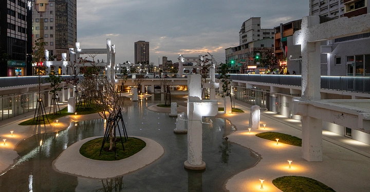 Galerie-Bild 5: Weiße Betonskulpturen stehen in einem beleuchteten Wasserbecken inmitten eines städtischen Platzes umgeben von hohen Gebäuden abends bei Dämmerung. Lampen und Pflanzen umgeben die Skulpturen.