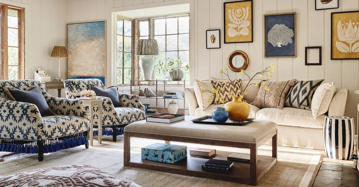 Sofa steht vor groβem Fenster Wohnzimmer dekoriert mit Kunstwerken und Pflanzen zwei Sessel mit blauem Muster und Beistelltischen beige Teppich und Holzmöbel verleihen Gemütlichkeit helle farbenfrohe Keramikvasen