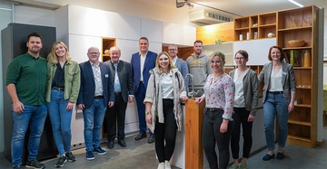 Mehrere Menschen stehen zusammen und lächeln Kamera in einer modernen Küchenausstellung. Sie tragen überwiegend legere Kleidung und stehen vor weißen Schränken und Holzregalen in einem gut beleuchteten Raum.