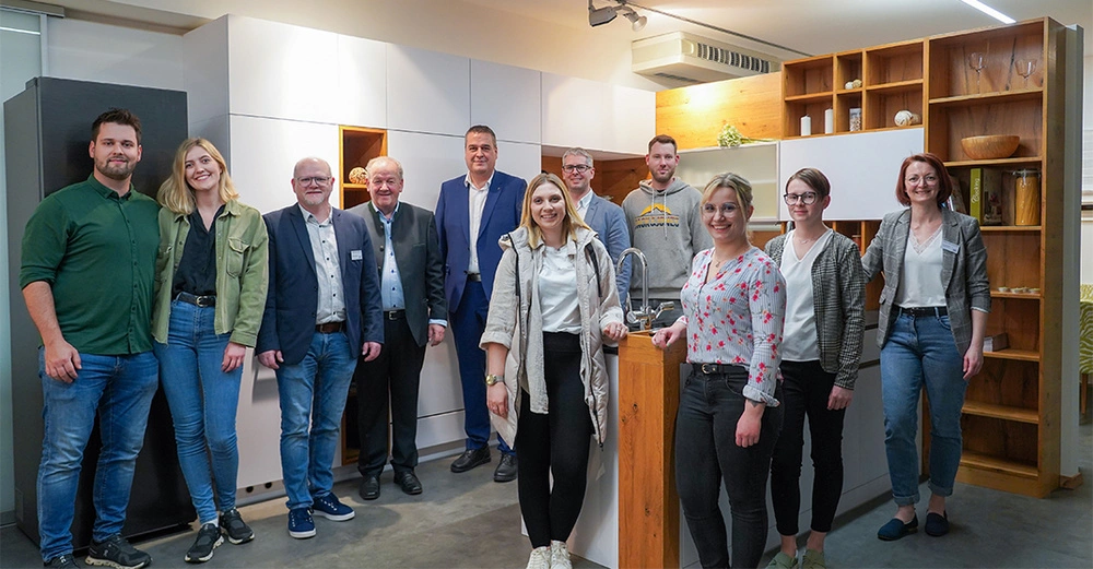 Mehrere Menschen stehen zusammen und lächeln Kamera in einer modernen Küchenausstellung. Sie tragen überwiegend legere Kleidung und stehen vor weißen Schränken und Holzregalen in einem gut beleuchteten Raum.