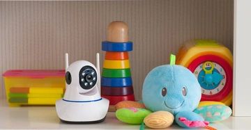 Babyphone mit Kamera steht neben einem Spielzeug-Okto, Stapelspiel und Lernuhr auf einem Regal in einem Kinderzimmer. Bücher sind auf dem oberen Regal zu sehen.